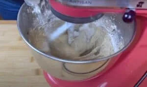 Red stand mixer kneading dough in a stainless steel bowl on a wooden surface.